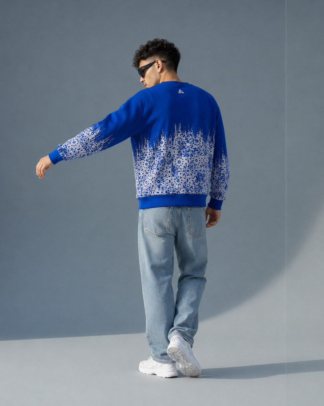 A man wearing a royal blue crewneck sweater with a white snowflake pattern on the lower half, paired with light blue jeans and white sneakers, shown from a back-three-quarter view against a gray studio background.