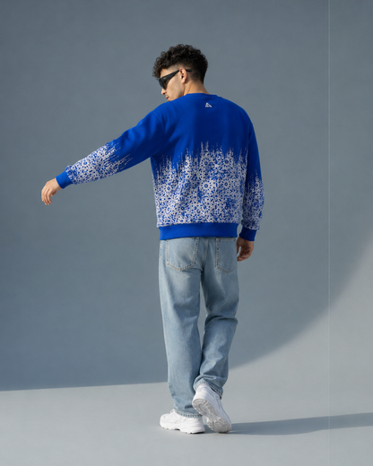 A man wearing a royal blue crewneck sweater with a white snowflake pattern on the lower half, paired with light blue jeans and white sneakers, shown from a back-three-quarter view against a gray studio background.