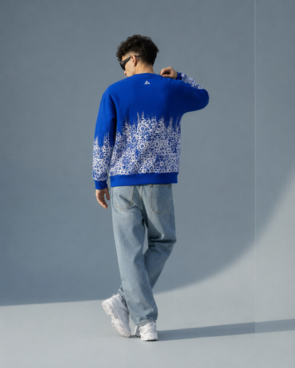 A man wearing a royal blue crewneck sweater with a white floral pattern on the lower half, paired with light blue jeans and white sneakers, shown from behind against a gray studio background.