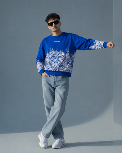 A man modeling a royal blue crewneck sweatshirt with a white forest tree pattern across the lower half and sleeves, paired with light blue jeans and white sneakers against a gray studio background.