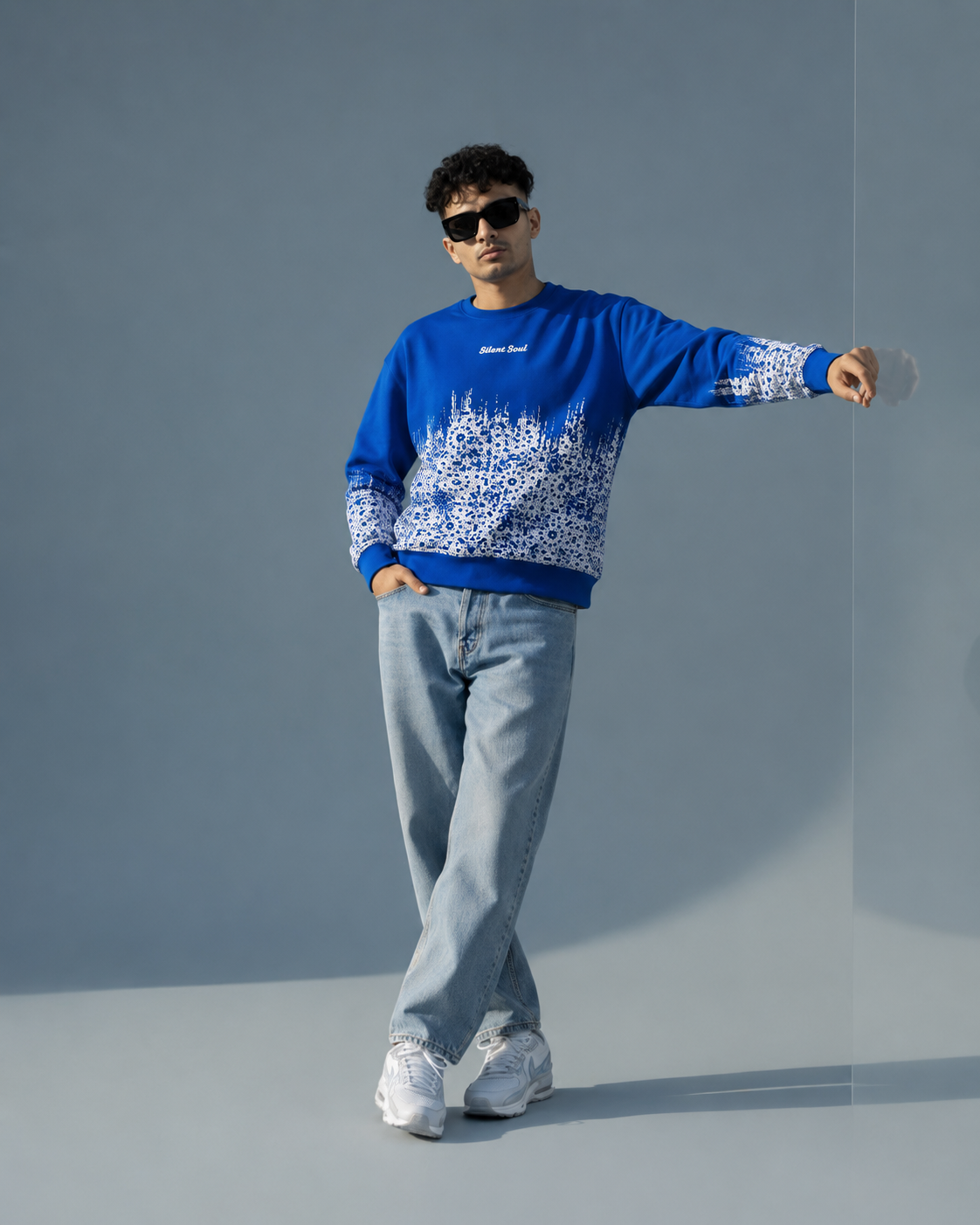 A man modeling a blue crewneck sweatshirt with a white graphic pattern on the lower half and sleeves, paired with light blue jeans and white sneakers, set against a minimalist gray studio background.