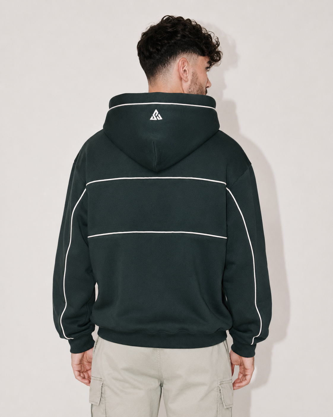 A man viewed from the back wearing a dark forest green hoodie with white contrast stitching and a small white geometric logo on the back of the neck, paired with light beige cargo pants against a plain light gray background.