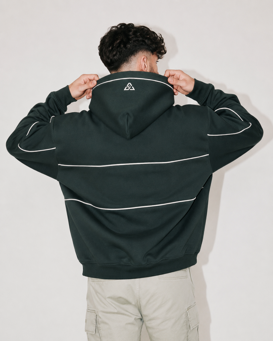 A man viewed from behind wearing a dark forest green hoodie with white contrast piping and a small white geometric logo on the back of the neck, paired with light beige cargo pants against a plain light gray background.