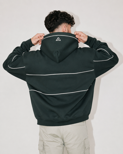 A man viewed from behind wearing a dark forest green hoodie with white contrast piping and a small white geometric logo on the back of the neck, paired with light beige cargo pants against a plain light gray background.