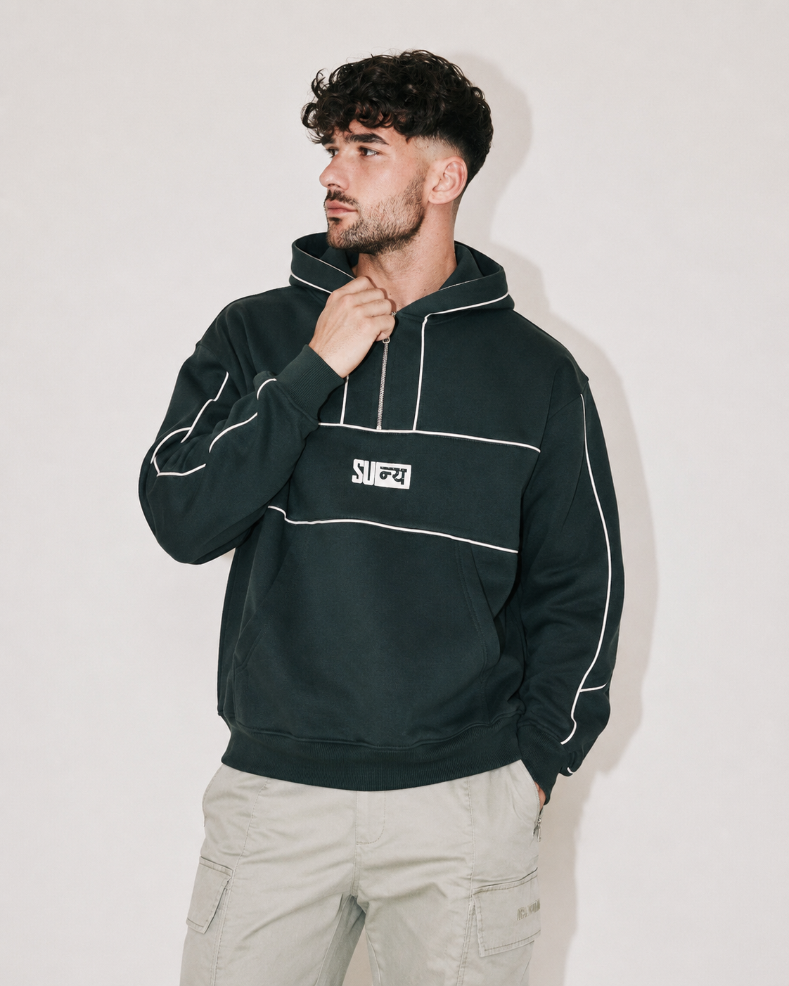 A man wearing a dark forest green pullover hoodie with white contrast stitching and a white chest patch logo, paired with light beige cargo pants, standing against a plain light gray wall.