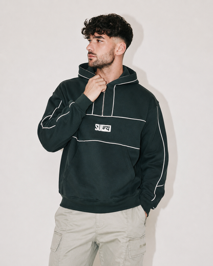 A man wearing a dark forest green pullover hoodie with white contrast stitching and a white chest patch logo, paired with light beige cargo pants, standing against a plain light gray wall.