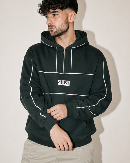 A man wearing a dark forest green half-zip hoodie with white contrast piping and a white chest logo, paired with light beige pants, set against a plain beige wall.