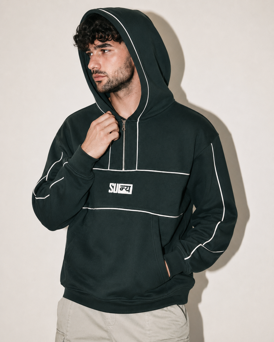 A man wearing a dark forest green pullover hoodie with white piping details and a white chest logo, paired with light beige pants, posing against a plain beige wall.