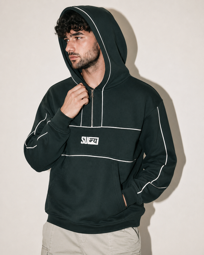 A man wearing a dark forest green pullover hoodie with white piping details and a white chest logo, paired with light beige pants, posing against a plain beige wall.