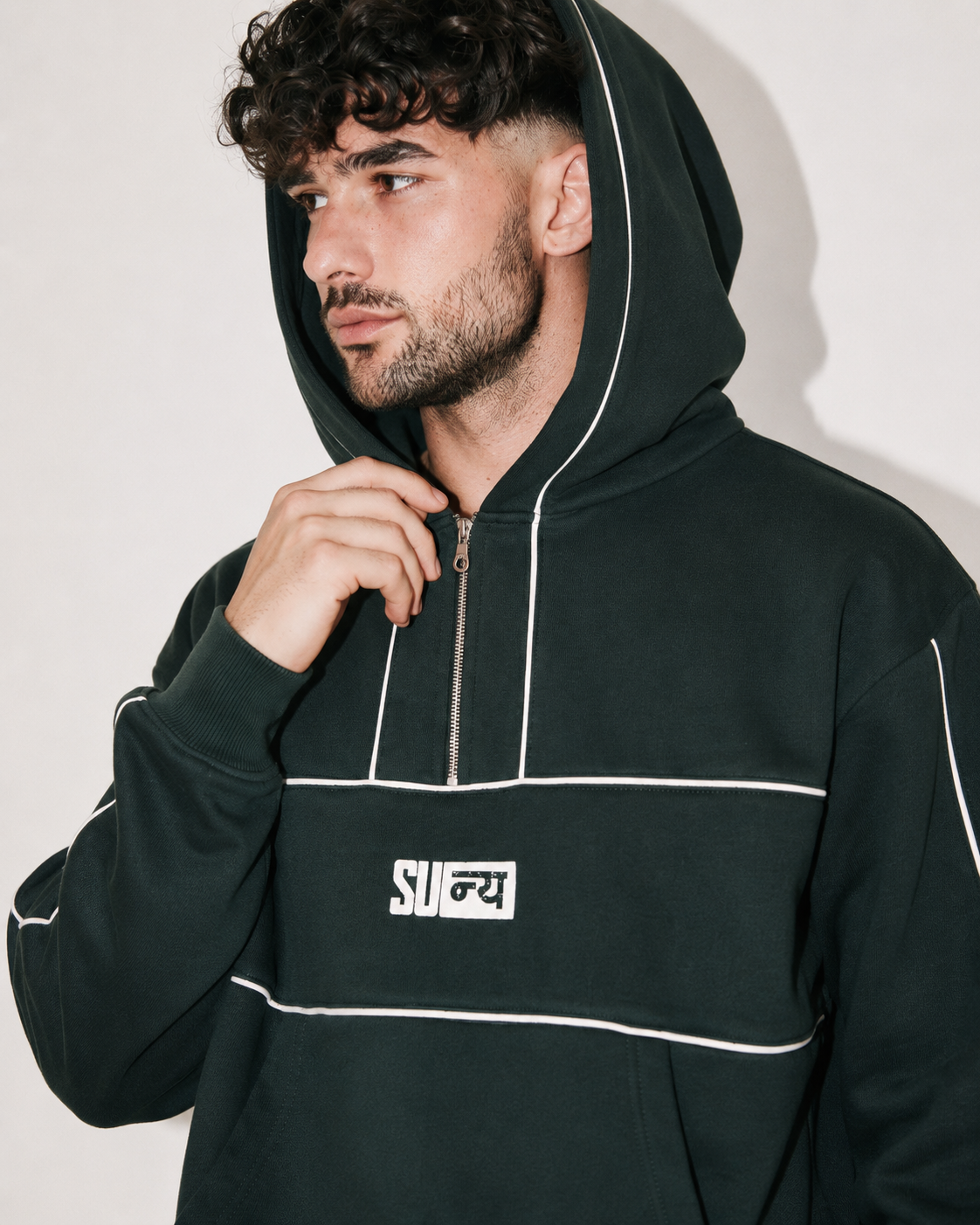 A man wearing a dark forest green pullover hoodie with white piping details and a white chest logo, posed against a light gray background.