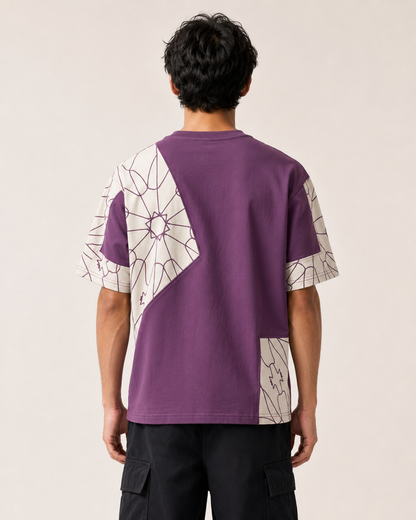 A man viewed from behind wearing a purple short-sleeve t-shirt with a white and black geometric spiderweb pattern on the back and sleeves, paired with black cargo pants, against a plain beige background.