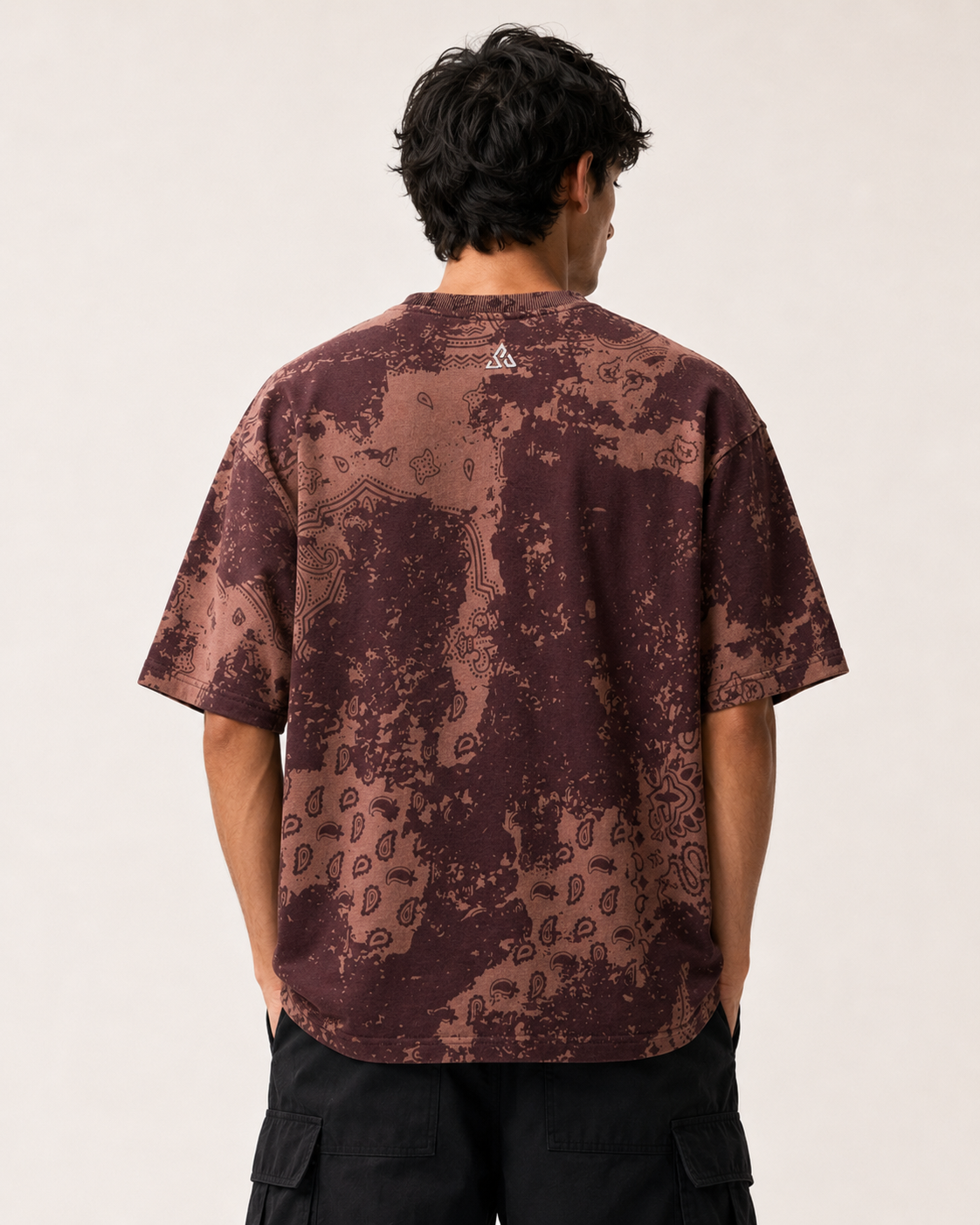 A man viewed from behind wearing a short-sleeve t-shirt with a brown and dark red abstract pattern, paired with black cargo pants, against a plain off-white background.