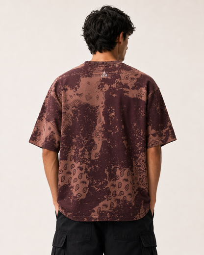 A man viewed from behind wearing a short-sleeve t-shirt with a brown and dark red abstract pattern, paired with black cargo pants, against a plain off-white background.