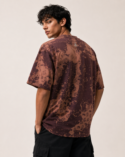 A man wearing a brown and dark purple abstract patterned t-shirt, shown from the back against a plain light beige background.
