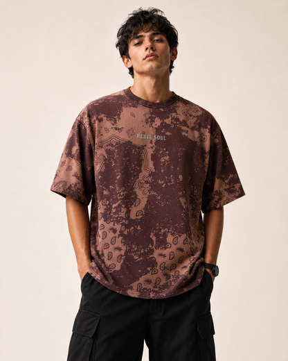 A man wearing a brown and dark brown paisley patterned t-shirt with 'RESILIENT SOUL' printed on the chest, paired with black cargo pants, against a plain beige background.
