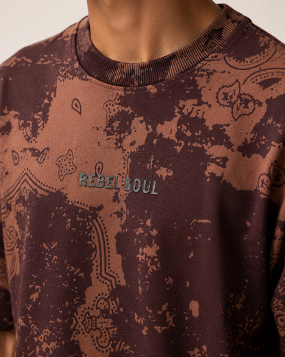 Close-up of a person wearing a short-sleeve crewneck sweatshirt with a brown and dark brown distressed paisley pattern and 'REBEL SOUL' embroidered on the chest.