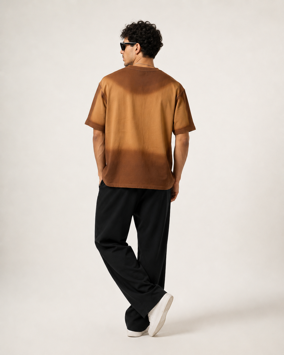 A man wearing a brown short-sleeve t-shirt with a gradient color-block design, black pants, and white sneakers, photographed from behind against a plain light background.