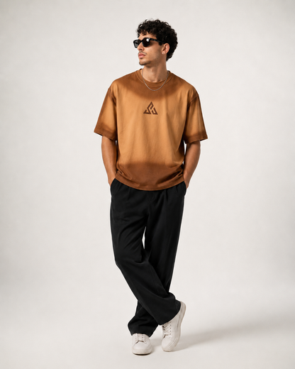 A man wearing a bronze-brown short-sleeve t-shirt with a small black geometric logo on the left chest, paired with black pants and white sneakers, standing against a light gray studio background.