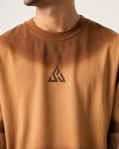 A close-up of a person wearing a brown crewneck sweatshirt with a dark geometric triangular logo on the chest, set against a plain light background.