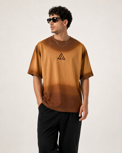A man wearing a brown gradient t-shirt with a black geometric logo on the chest, paired with black pants and sunglasses, standing against a plain light background.