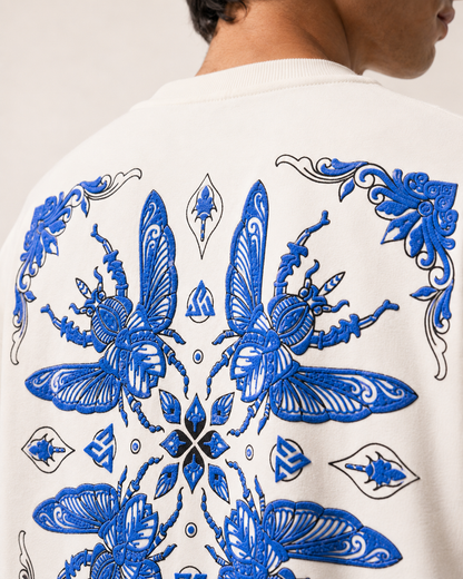 Close-up of the back of a white crewneck sweatshirt featuring a large, detailed blue embroidered butterfly and floral pattern, worn by a model.