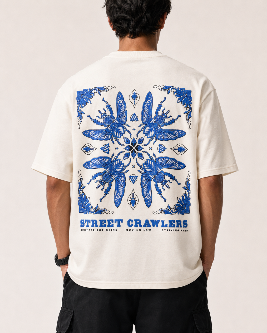A man viewed from behind wearing a cream-colored t-shirt with a large blue mandala-style graphic and 'STREET CRAWLERS' text printed on the back, paired with black cargo pants against a light beige studio background.