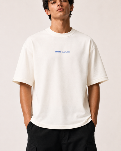 A man wearing an oversized off-white crewneck t-shirt with a small blue 'STREET CLOTHING' logo on the chest, paired with black cargo pants, against a solid light beige background.