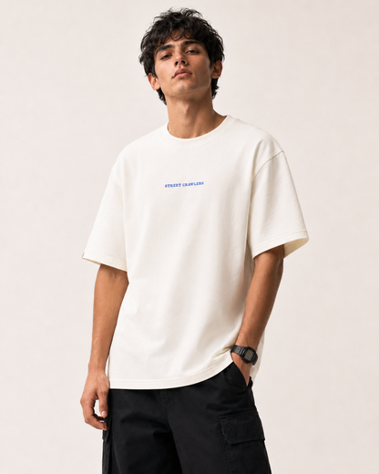 A man wearing a cream-colored short-sleeve t-shirt with a small blue 'STREET CARAVAN' logo on the chest, paired with black cargo pants, against a plain light beige background.