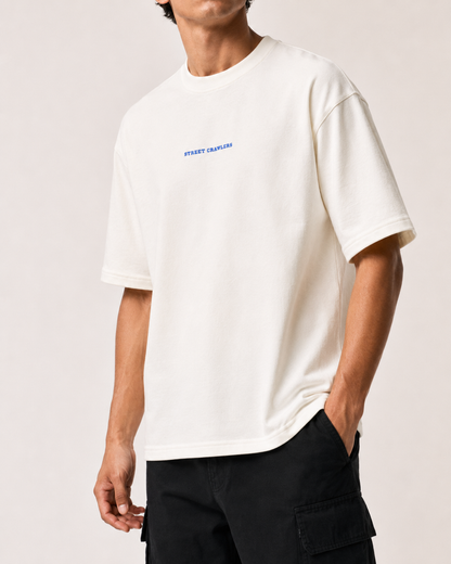 A man wearing a cream-colored short-sleeve t-shirt with 'STREET CLOTHING' printed in blue on the chest, paired with black cargo pants, against a light beige background.