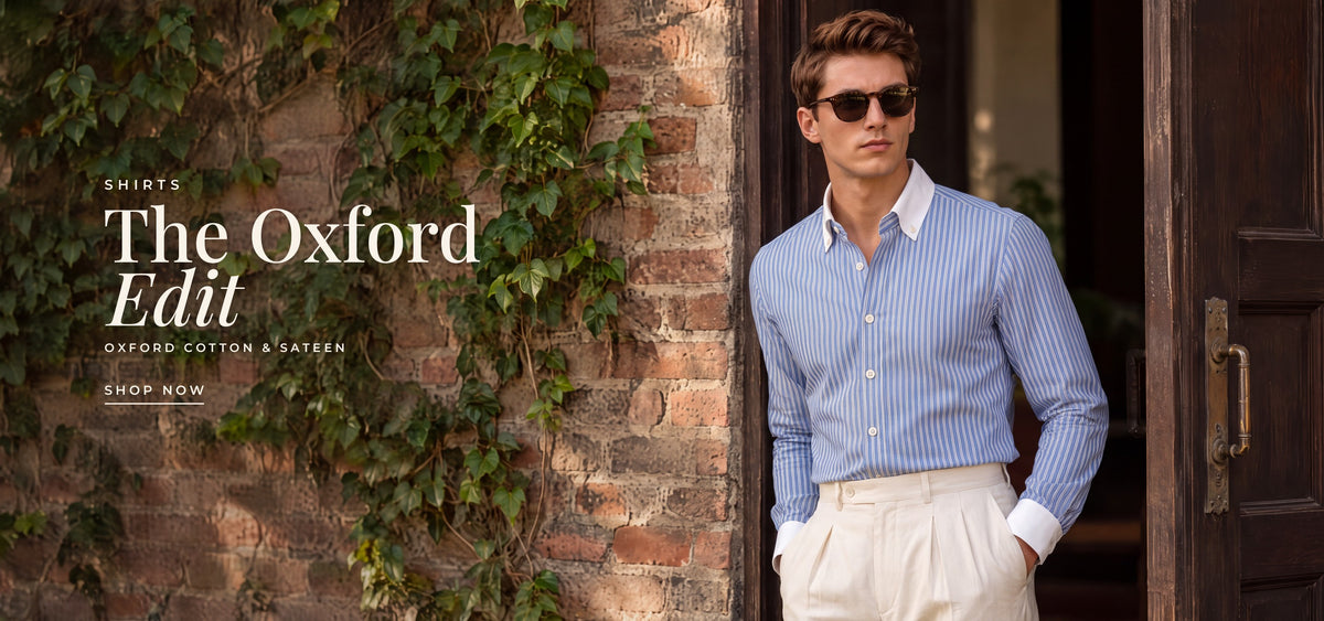 Shirts: The Oxford Edit, Oxford cotton and sateen. A man in the Blue Winchester shirt and tortoiseshell sunglasses by a brick wall and an open wooden door. Shop shirts.