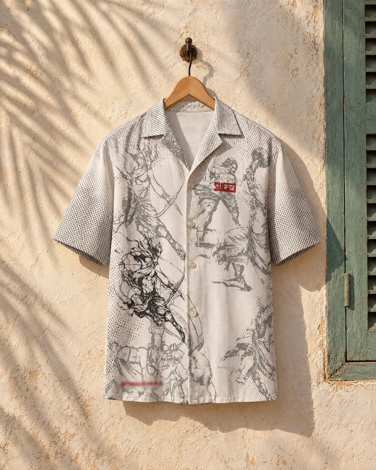 MOKSHA shirt hung on a wooden hanger against a sunlit whitewashed wall beside a green shutter