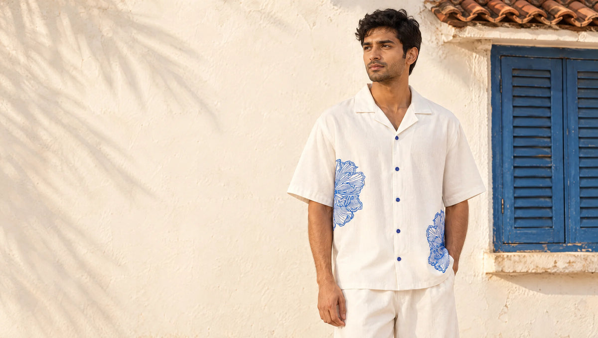 Man in the bone-white Vinelet camp-collar shirt with cobalt embroidered florals, standing by a whitewashed Goan wall and blue shutters in afternoon light