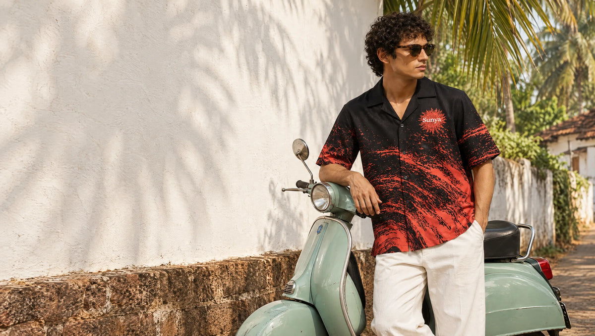 Man in the black BLAZE shirt with red splatter wave and Sunya sun emblem, leaning on a mint scooter in a Goan lane