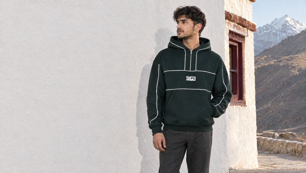 A young man in the forest-green NEXUS half-zip hoodie against a whitewashed monastery wall in Spiti