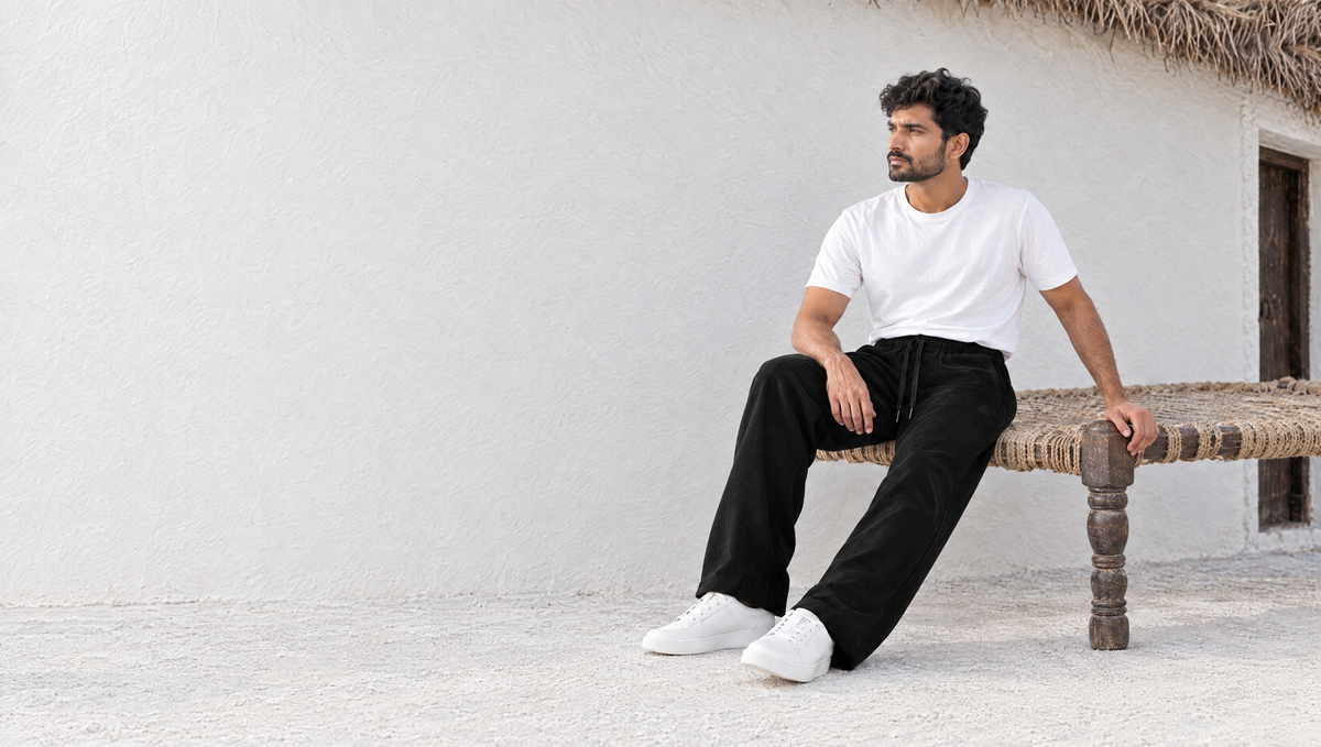 The TAMAS cord joggers in black, worn seated on a rope charpai outside a whitewashed Kutch bhunga