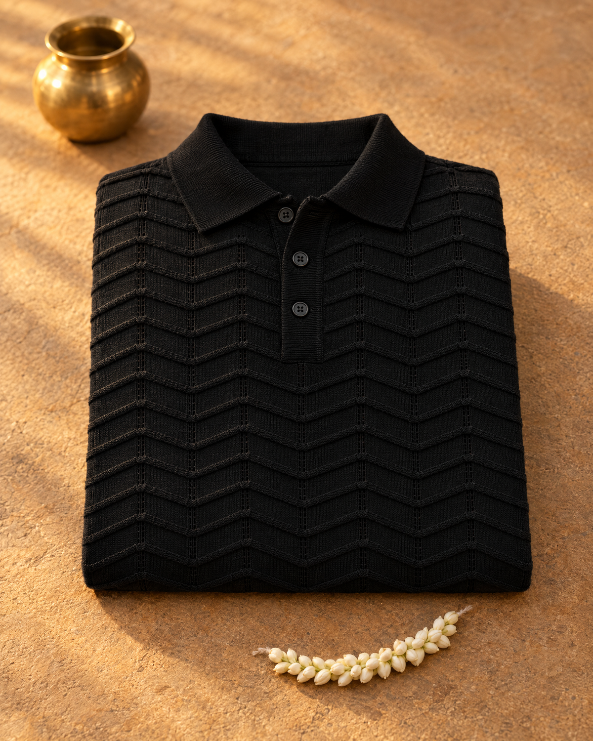 The black FERN knit polo folded on warm sandstone beside jasmine and a brass lota