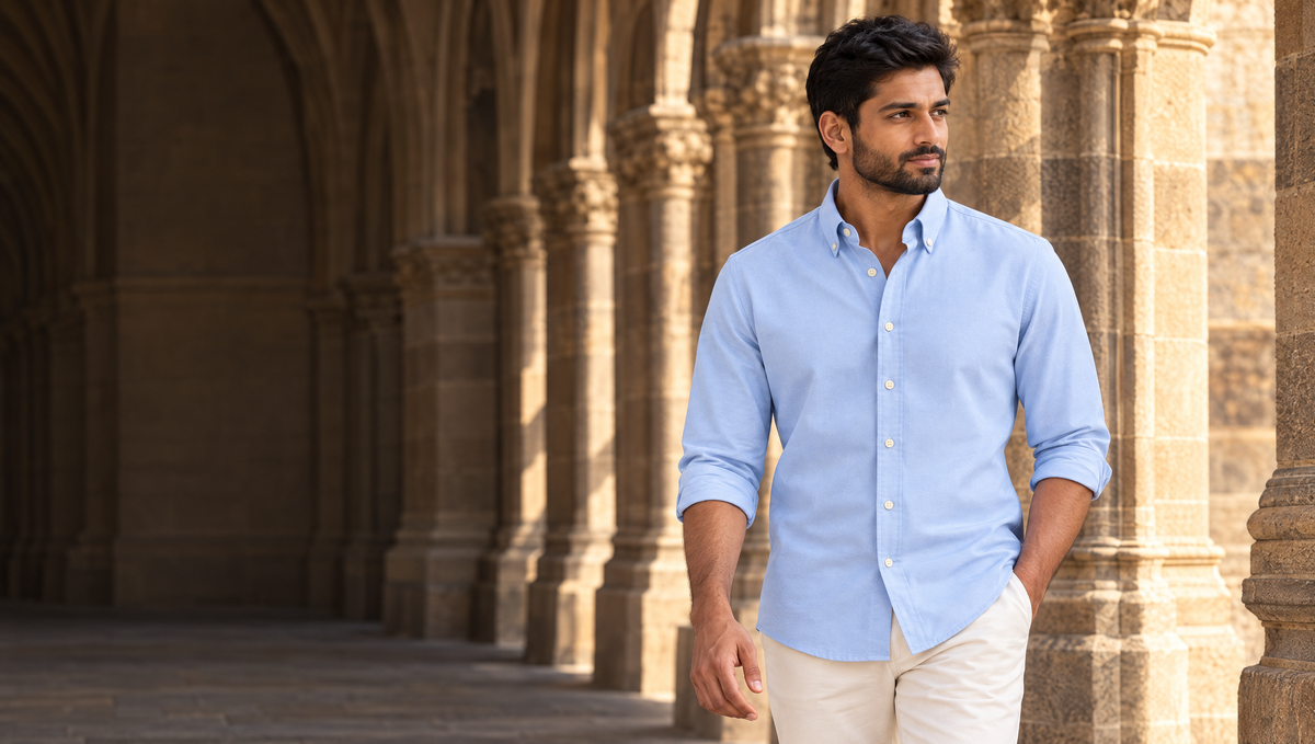 A man in the Powder Oxford Shirt, sleeves rolled and collar points buttoned down, walking through a Gothic stone arcade in Bombay