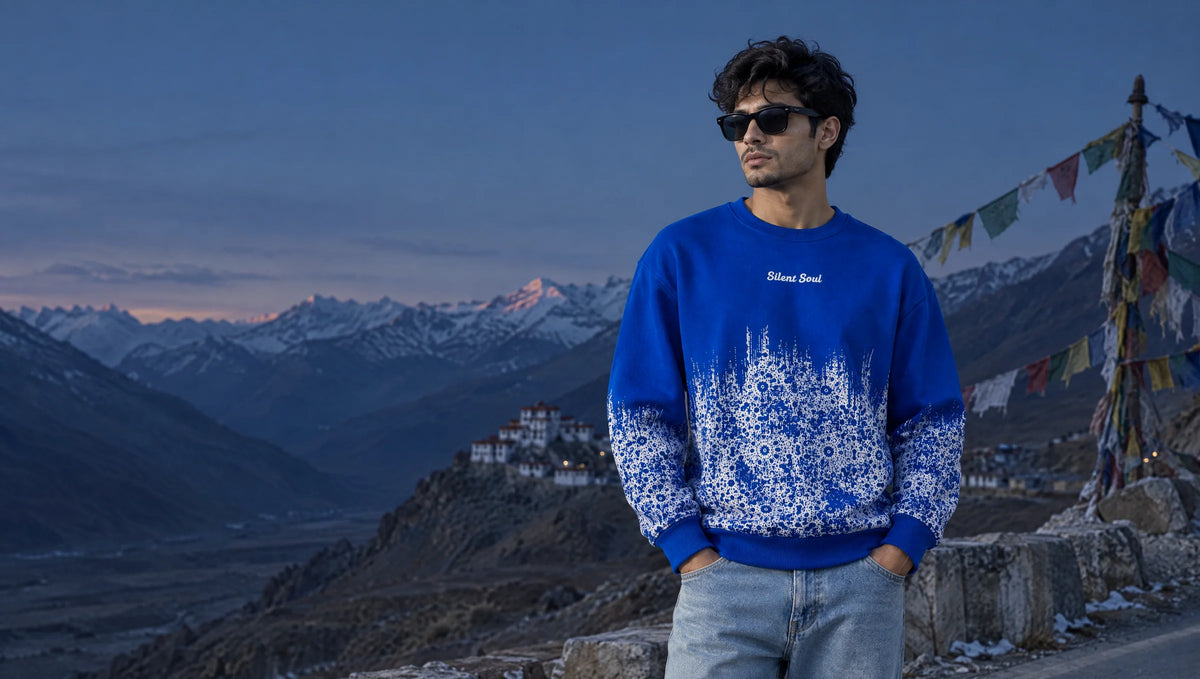 Man in a royal-blue MAUN sweatshirt with white floral rising from the hem, on a Spiti mountain road at blue dusk with prayer flags and a hilltop monastery behind