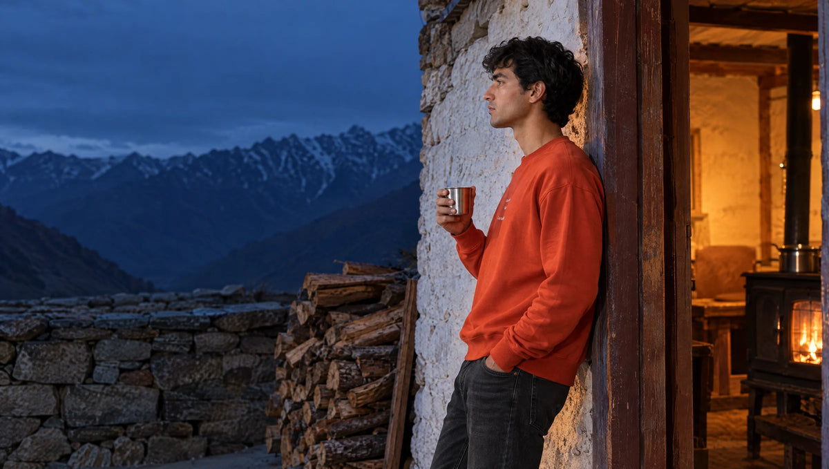 Man in a rust-orange Nor Memories sweatshirt leaning in a homestay doorway with a cup of chai, wood stove glowing inside, blue dusk over the mountains