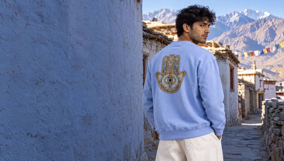 Man seen from behind in a sky-blue Vibrant Hamsa sweatshirt, the multicolour Hamsa on his back, in a whitewashed Ladakhi village lane on a crisp morning