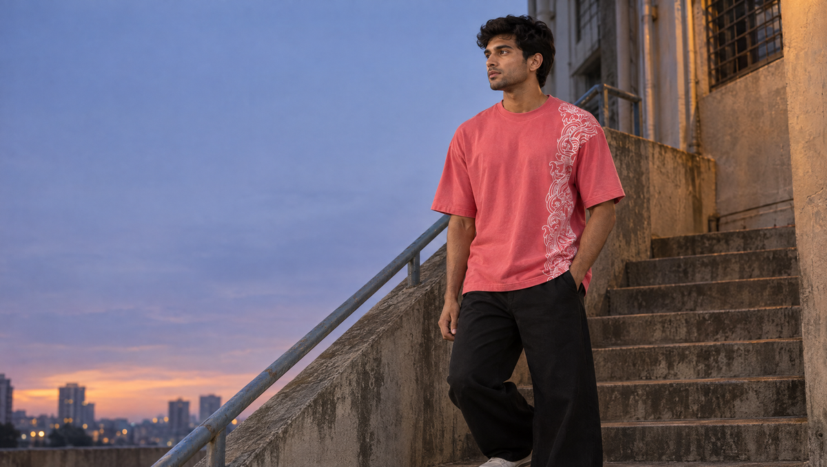 A man in the coral-red Ornate Cosmos tee, white scrollwork running down one side, on a concrete stairway in Mumbai at dusk