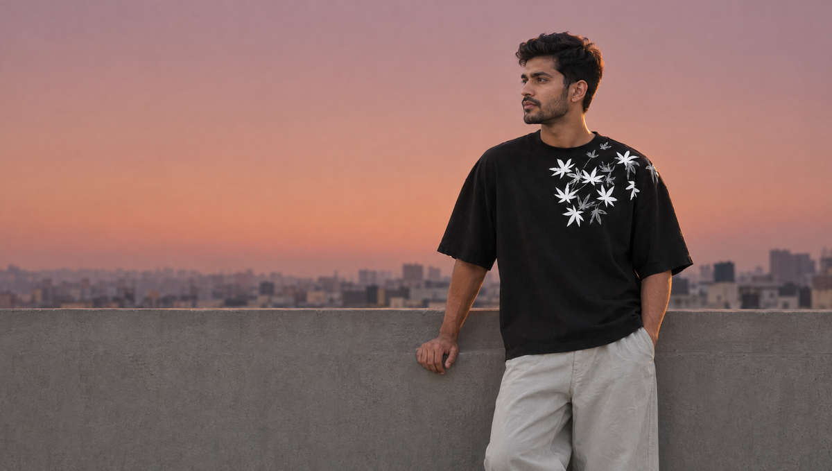 A man in the black Botanical Serenity tee, white maple leaves across the chest and shoulder, on a Mumbai rooftop at sunset