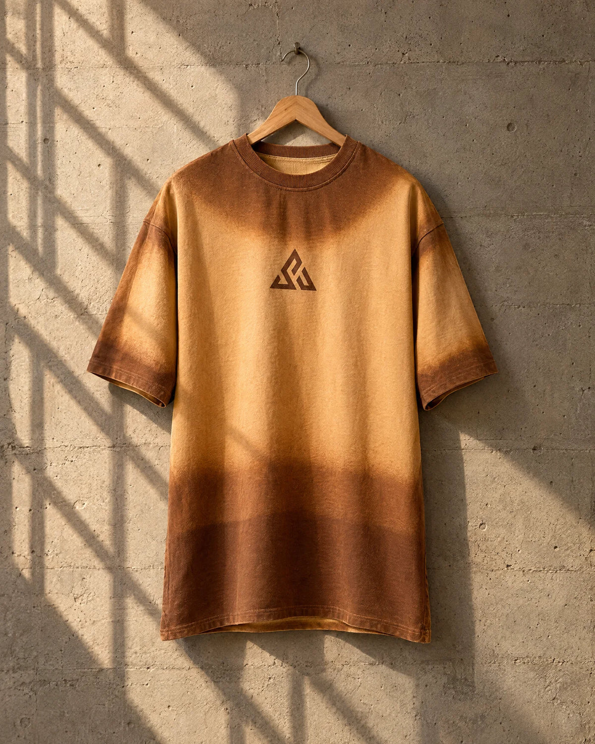 Sand-to-brown tee on a wooden hanger against a concrete wall striped with window-grille shadows