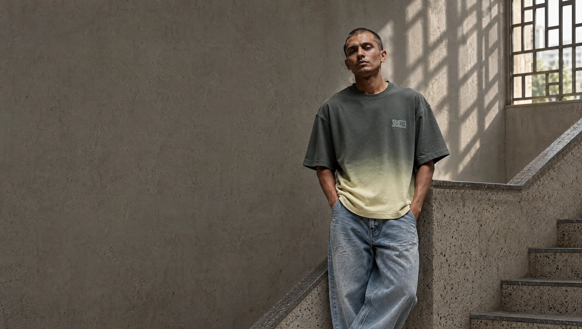 Man in an olive-to-sage ombré tee and light denim leaning on a terrazzo stair rail under window-grille shadows
