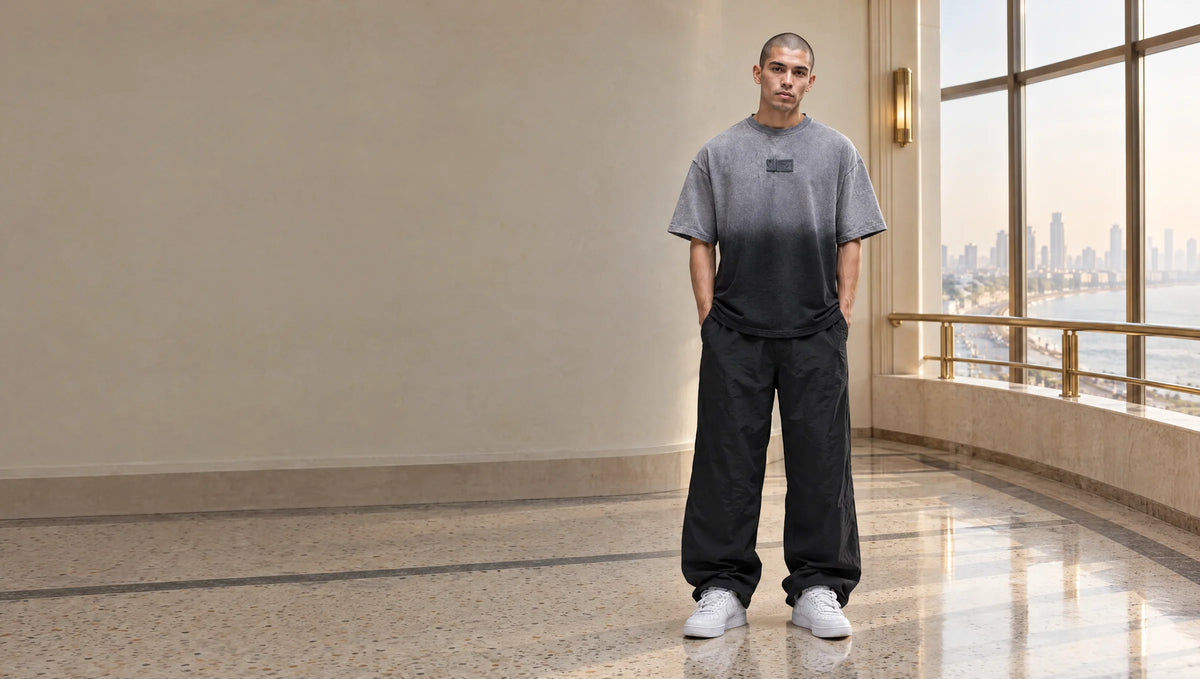 Man in an oversized tee fading from ash-grey to charcoal-black, standing in an empty art-deco lobby overlooking Marine Drive
