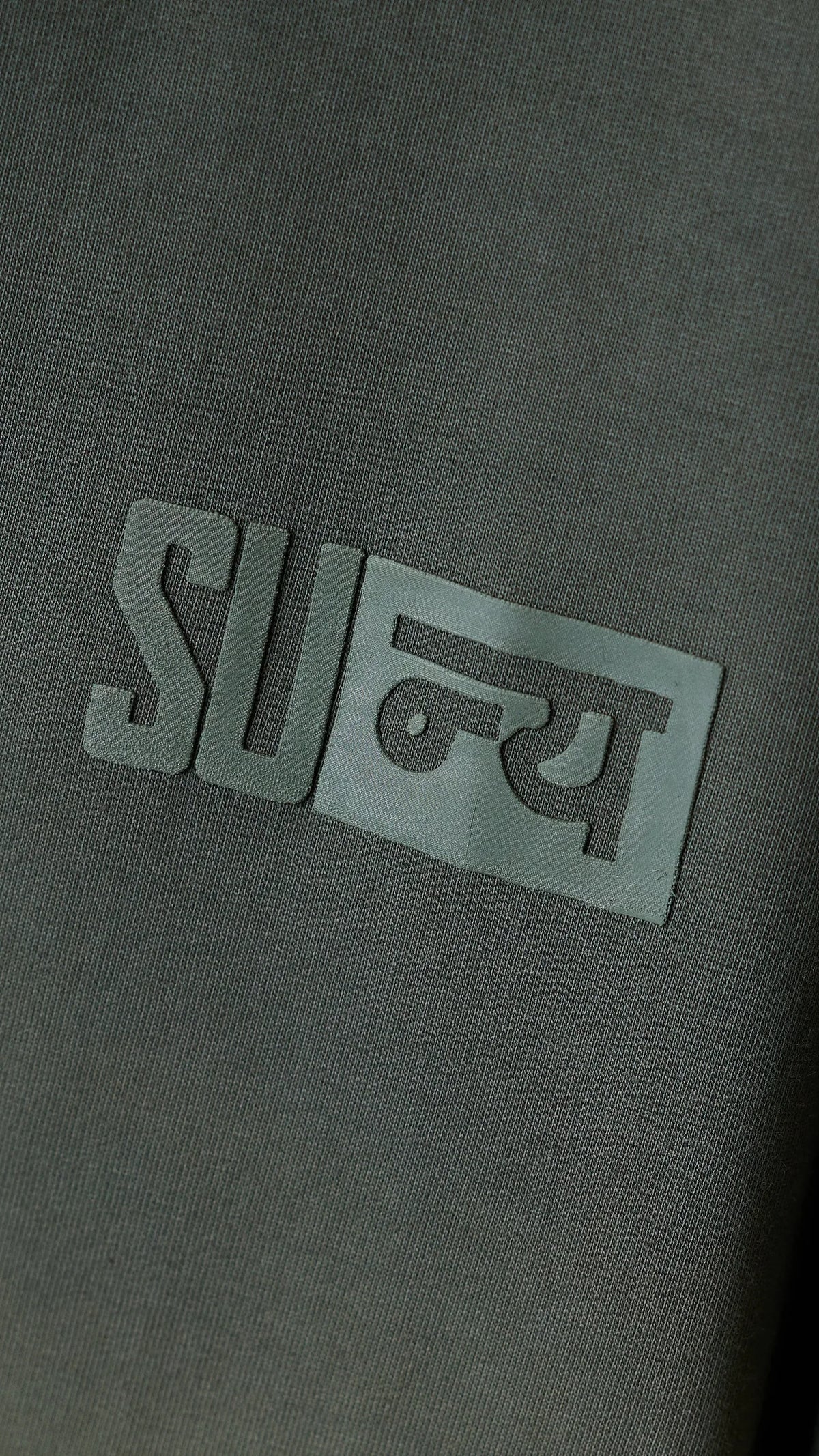 Close-up of the raised green-grey boxed Sunya badge on olive cotton