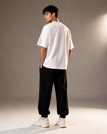 A man wearing a white oversized short-sleeve t-shirt, black jogger pants, and white sneakers, shown from a rear three-quarter angle against a neutral gray studio background.