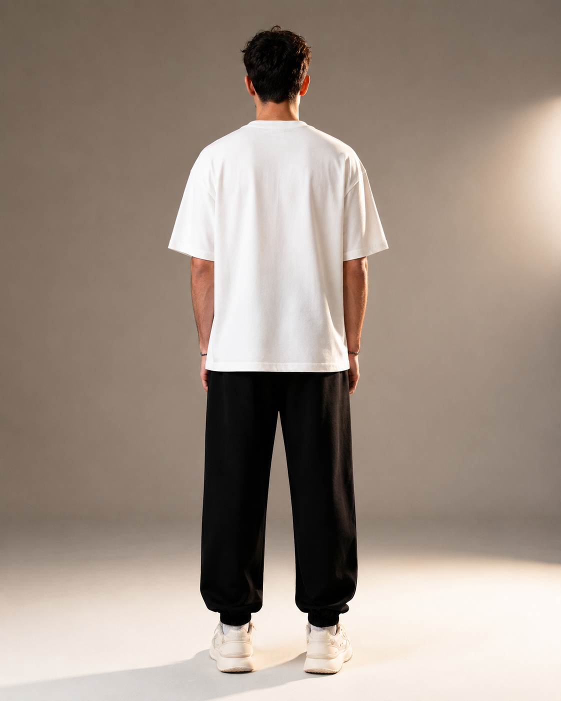 A man viewed from behind wearing a white short-sleeve t-shirt, black baggy sweatpants, and white sneakers, standing against a neutral gray studio background.