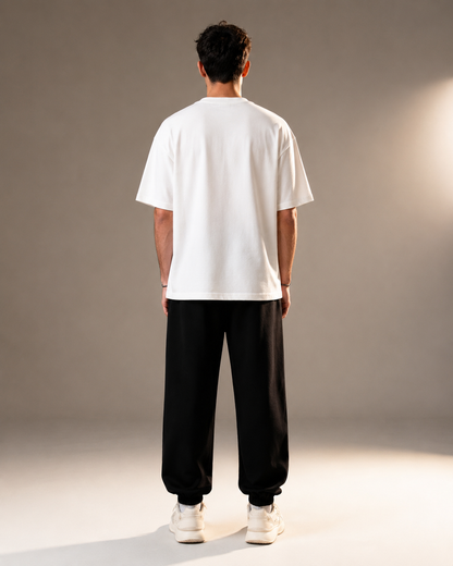 A man viewed from behind wearing a white short-sleeve t-shirt, black baggy sweatpants, and white sneakers, standing against a neutral gray studio background.