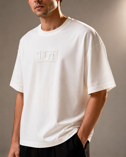 A man wearing an oversized white short-sleeve t-shirt with a tonal embossed Gucci logo box on the chest, paired with black trousers, against a neutral gray studio background.
